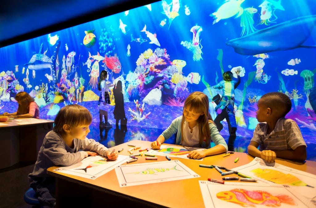 Kunst auf der Kinderbiennale: Kinder malen an Tisch, dahinter wird ein Aquarium an die Wand projiziert