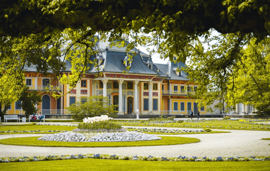 Frühlingsblumen blühen in den gepflegten Beeten vor dem Wasserpalais von Schloss Pillnitz in Dresden. Grüne Bäume rahmen die historische Fassade ein.