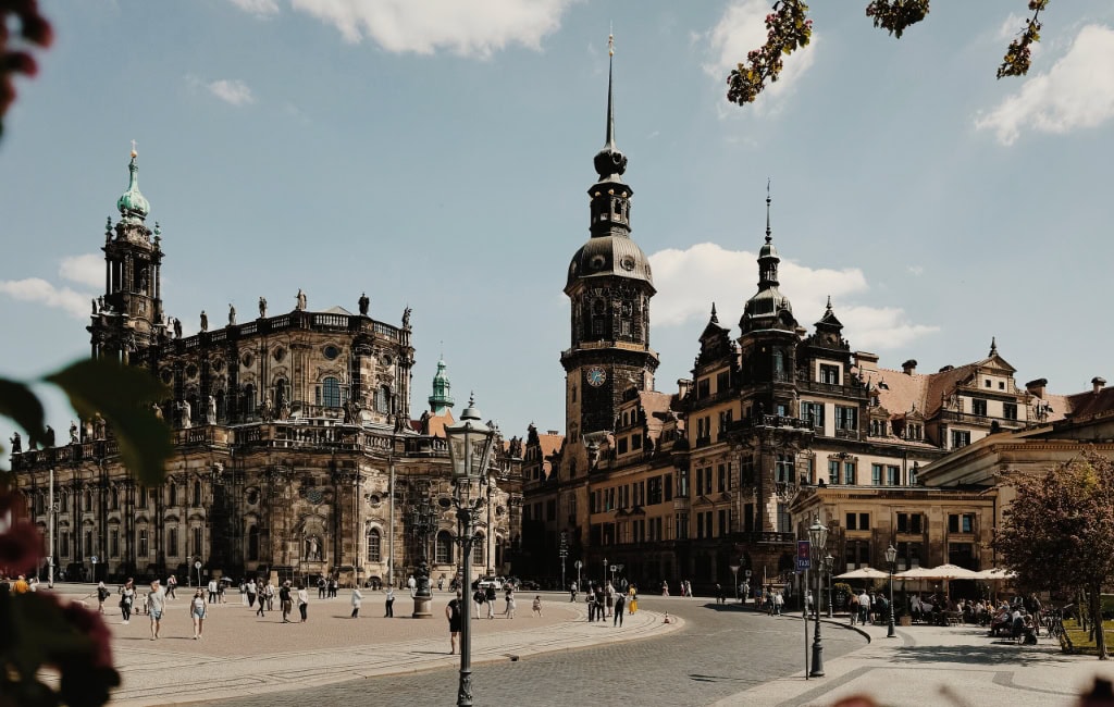 Blick vom Theaterplatz auf die Dresdner Hofkirche und das Residenzschloss mit dem markanten Hausmannsturm. Menschen spazieren über den Platz.