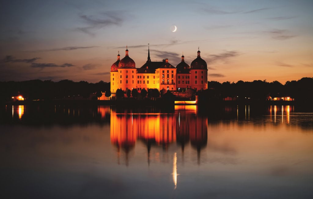 Das orange beleuchtete Barockschloss Moritzburg spiegelt sich bei Sonnenuntergang im ruhigen Schlossteich. Am Himmel steht eine schmale Mondsichel.