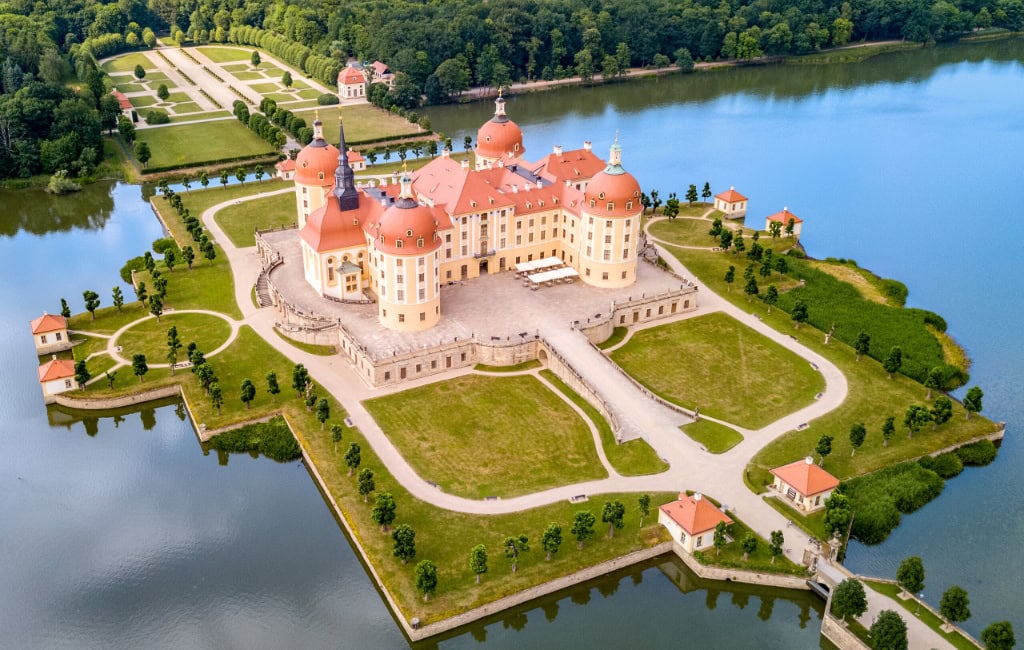 Luftaufnahme von Schloss Moritzburg auf einer künstlichen Insel inmitten eines Sees. Zu sehen sind die vier runden Ecktürme, die Zufahrtsbrücke und der barocke Schlossgarten.