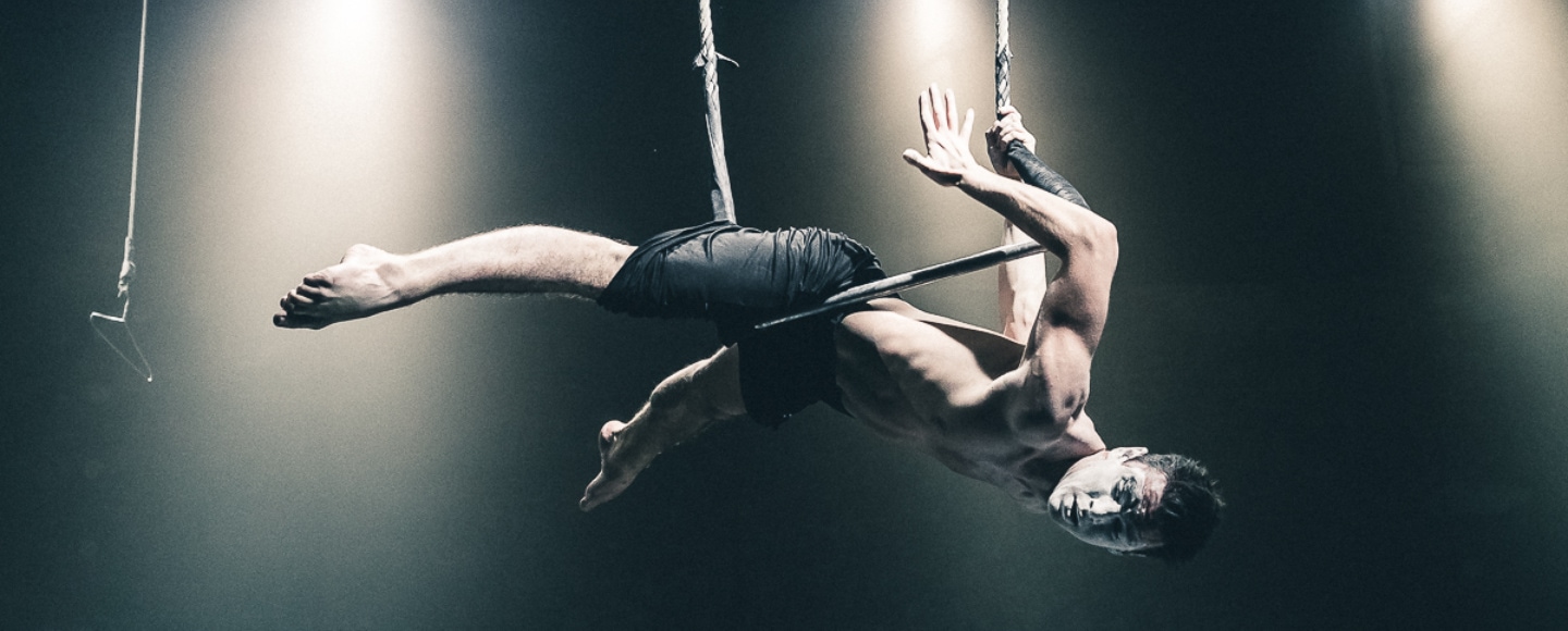 Ein Akrobat hängt horizontal an Seilen in der Luft und schaut in die Kamera. Sein Gesicht ist geschminkt.
