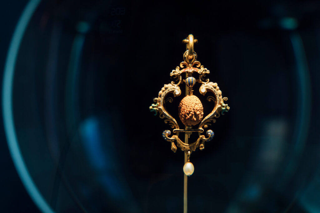 Goldener Anhänger mit filigranem Ornamentrahmen und geschnitztem Kirschkern vor dunklem blauem Hintergrund.