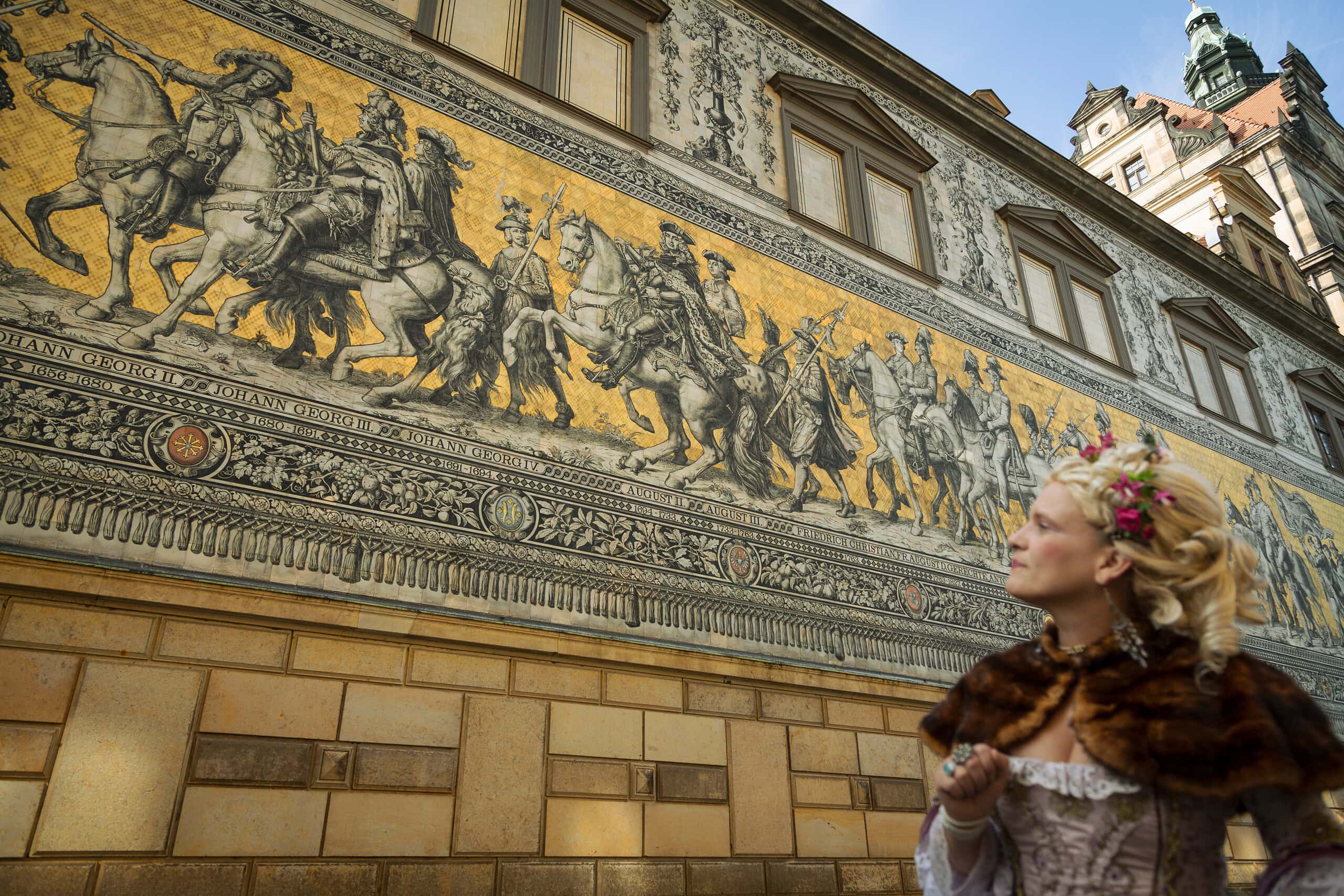 Residenzschloss Dresden: Fürstenzug an der Schlossfassade mit Reiterzug aus Meißner Porzellan. Davor steht eine Frau im barockem Kleid.