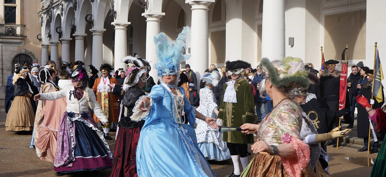 Mehrere Teilnehmerinnen und Teilnemer des Elbvenezianischen Carnevals in Dresden tanzen gemeinsam in farbenfohen, kunstvollen Kleidern und Masken im Barock-Stil.