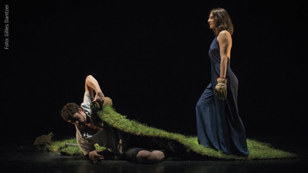 Mann kriecht unter Rasenrolle hervor, Frau in blauem Kleid steht daneben