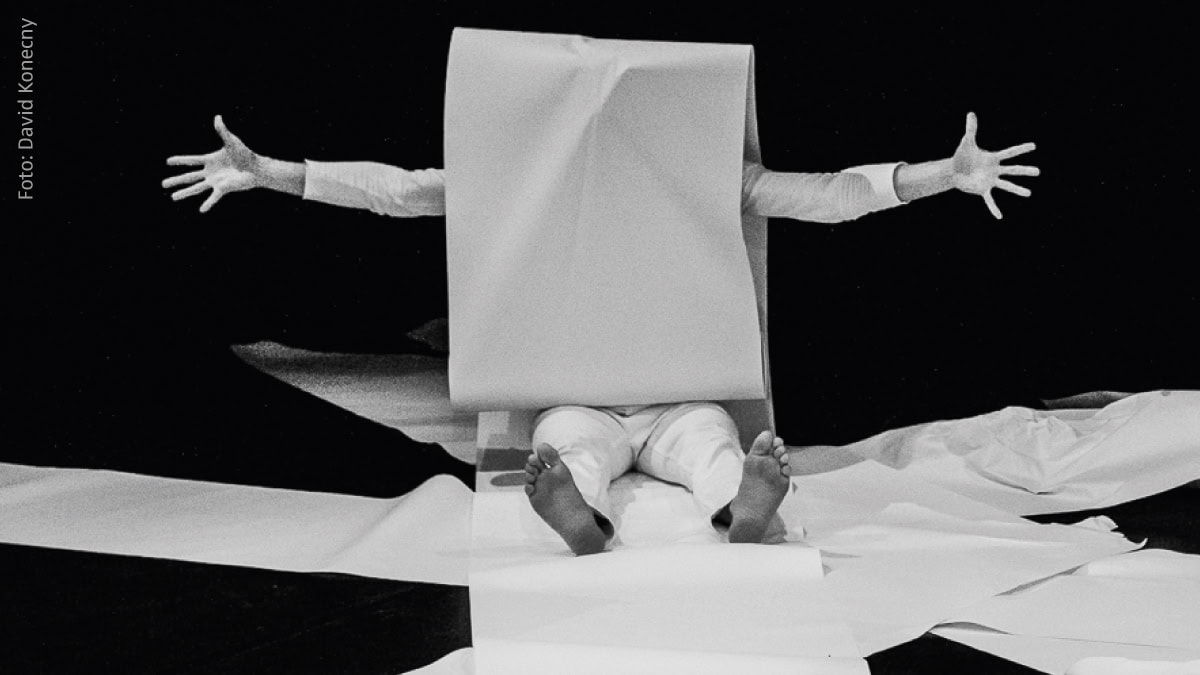Schwarz-Weiß-Bild einer Person, die unter einem großen Blatt Papier sitzt, Arme ausgestreckt