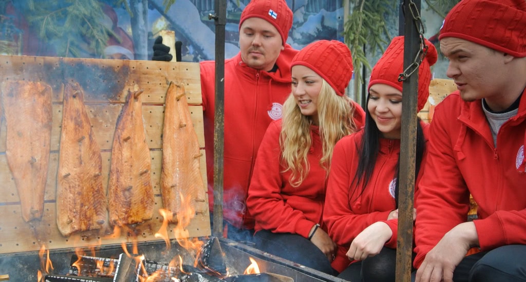 Zu sehen sind vier Teammitglieder des finnischen Weihnachtsmarkts, die um ein Feuer herum stehen, an dem gerade Flammlachs zuberreitet wird.