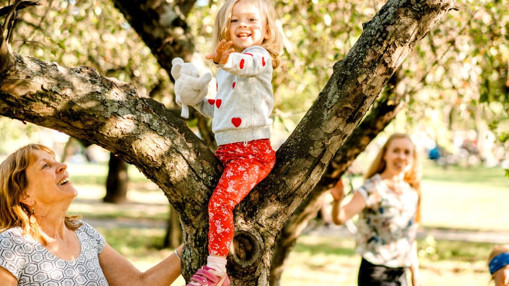 Ein kleines Kind sitzt lächelnd auf einem Ast in einem blühenden Baum, während zwei Erwachsene und ein weiteres Kind darunter im Park stehen und nach oben schauen.