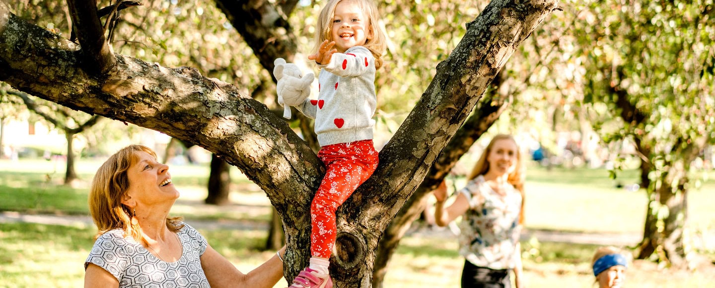 Ein kleines Kind sitzt lächelnd auf einem Ast in einem blühenden Baum, während zwei Erwachsene und ein weiteres Kind darunter im Park stehen und nach oben schauen.