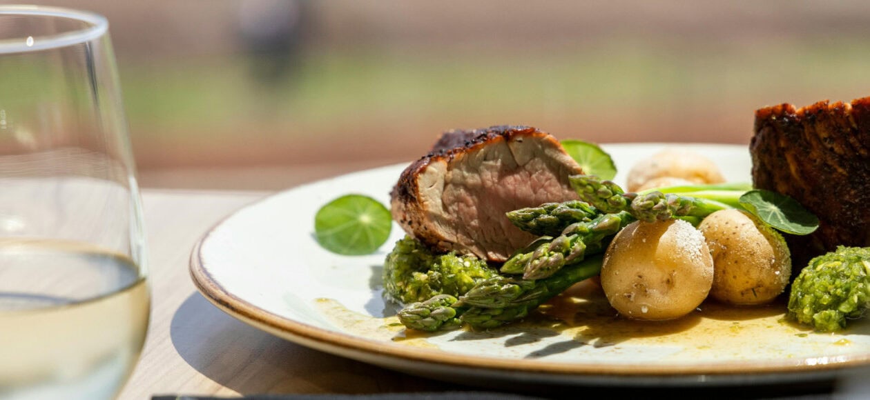 Teller mit rosa gebratenem Fleisch, grünem Spargel und jungen Kartoffeln mit Kräuterpesto; vorn ein Glas Weißwein, draußen im Sonnenlicht.