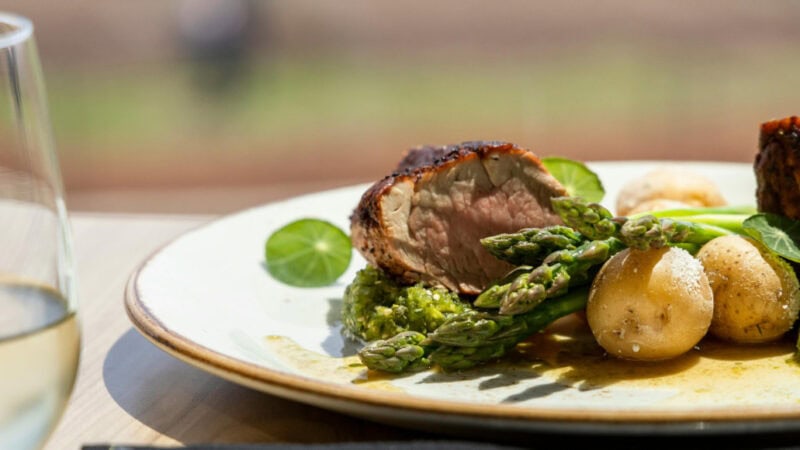 Teller mit rosa gebratenem Fleisch, grünem Spargel und jungen Kartoffeln mit Kräuterpesto; vorn ein Glas Weißwein, draußen im Sonnenlicht.
