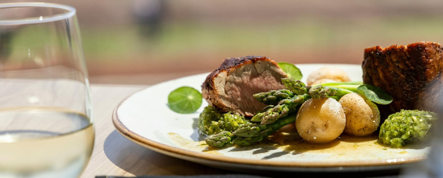 Teller mit rosa gebratenem Fleisch, grünem Spargel und jungen Kartoffeln mit Kräuterpesto; vorn ein Glas Weißwein, draußen im Sonnenlicht.