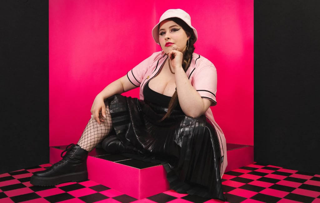 Eine Person in rosa-schwarzem Outfit und hellem Hut sitzt vor einer pinkfarbenen Kulisse mit schwarz-pink kariertem Boden.
