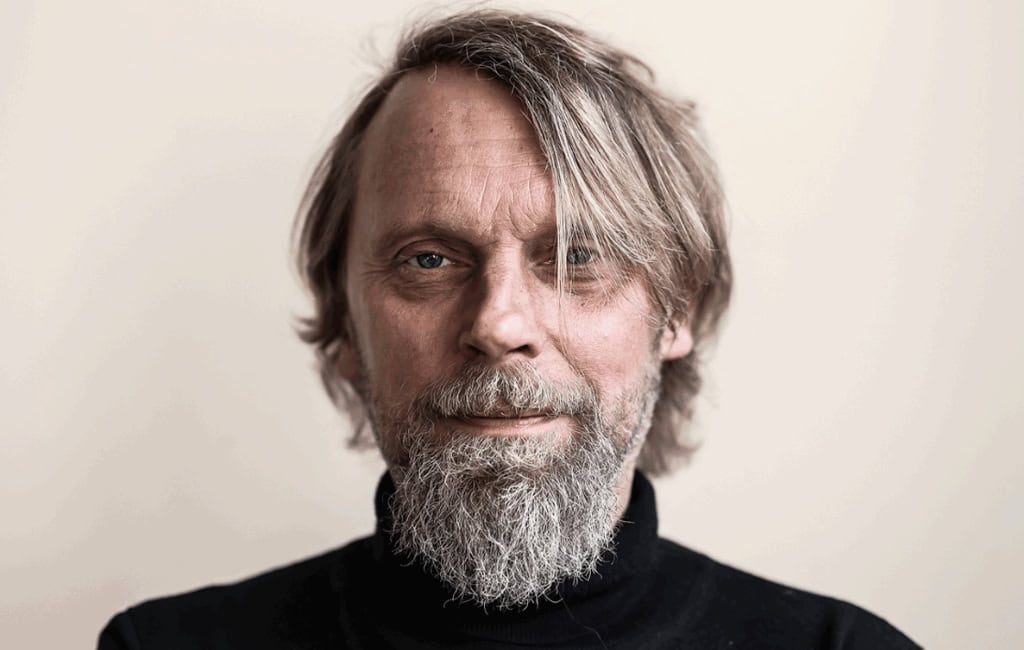 Porträt von Jens Balzer mit grauen Haaren und Vollbart und schwarzem Rollkragenpullover vor hellem Hintergrund.