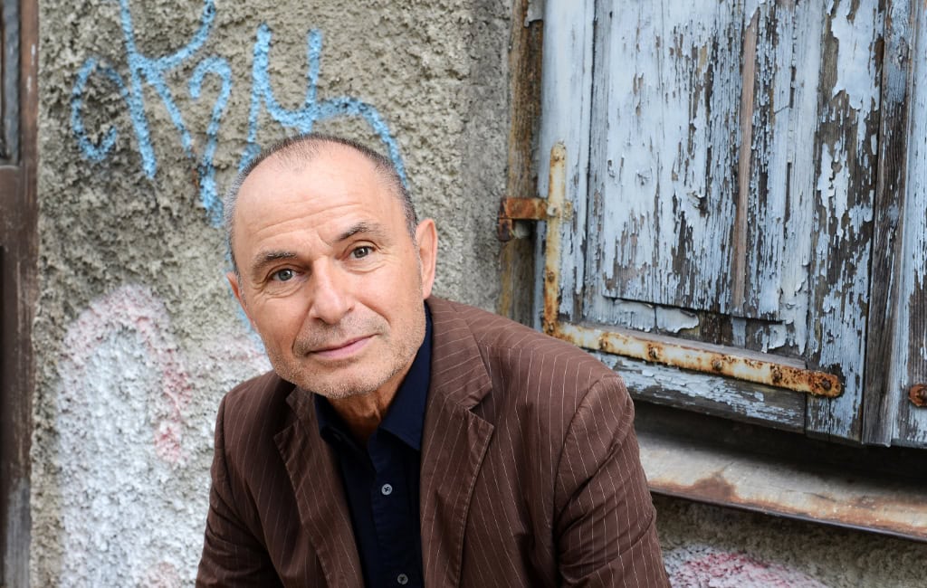 Autor Norbert Gstrein sitzt vor einer verwitterten Hauswand mit Graffiti und blickt in die Kamera.