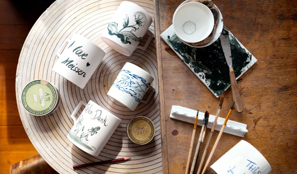 Verschiedene Tassen, Pinsel und Farben auf Holztisch in einer Porzellanmalerei-Werkstatt