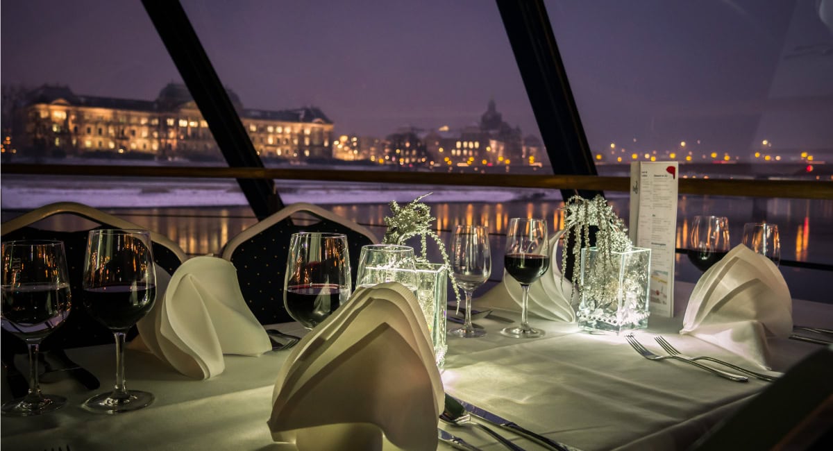 Festlich gedeckter Tisch mit gefalteten weißen Servietten, Rotweingläsern und leuchtender Deko auf einem Schiff derWeißen Flotte Sachsens; durch die Fenster sieht man abends die beleuchtete Dresdner Skyline und Lichtreflexe auf der Elbe.