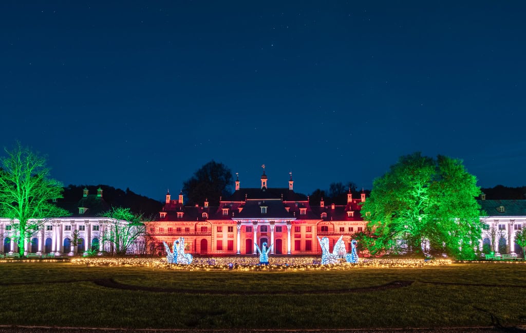 Nachtsicht auf Schloss Pillnitz in Dresden: rot illuminierte Fassade, links und rechts grün angestrahlte Bäume, im Vordergrund funkelnde Licht-Schwäne und Lichterketten unter klarem Sternenhimmel.