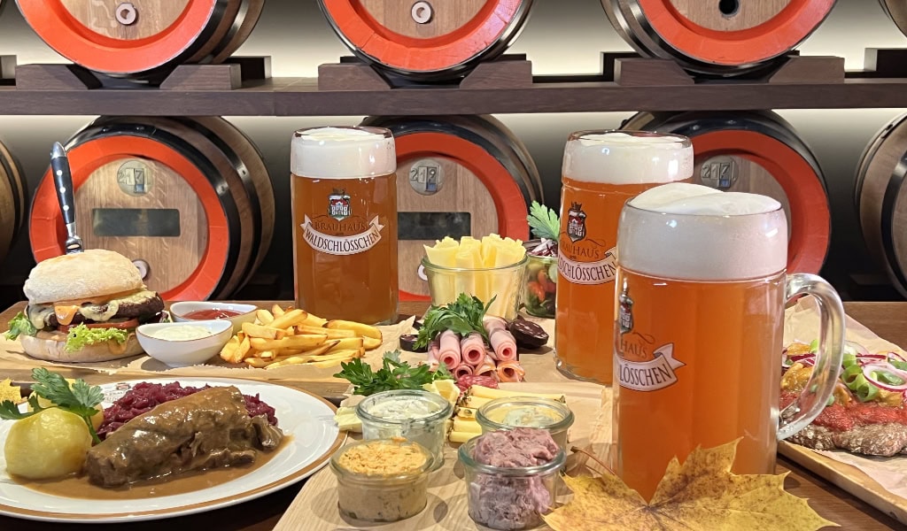 Rustikale Brauhaus-Tafel mit drei großen Bierkrügen, deftigen Speisen und Holzfässern im Hintergrund dicht aufgereiht. 