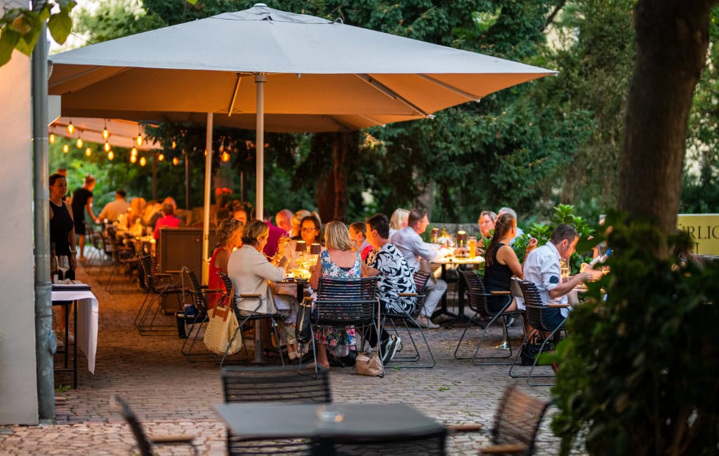 Innenhof von Mario Pattis Restaurant mit Bäumen und Gästen unter Sonnenschirm 