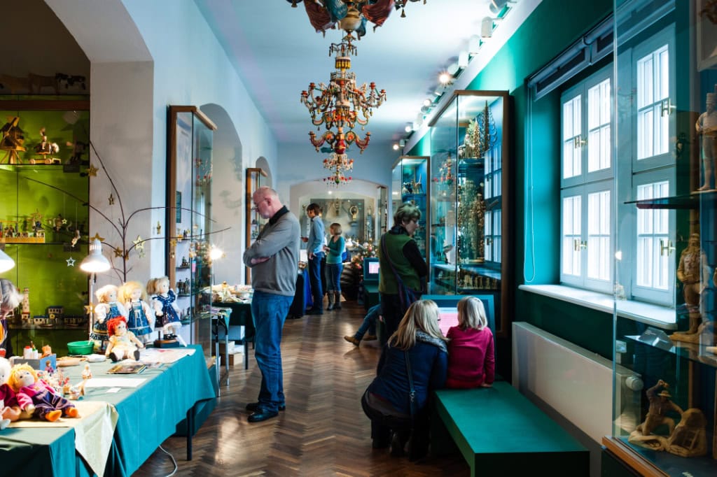 Gezeigt wird der Flur einer Ausstellung. Mehrere Menschen schauen sich die verschiedenen Gegenstände an.