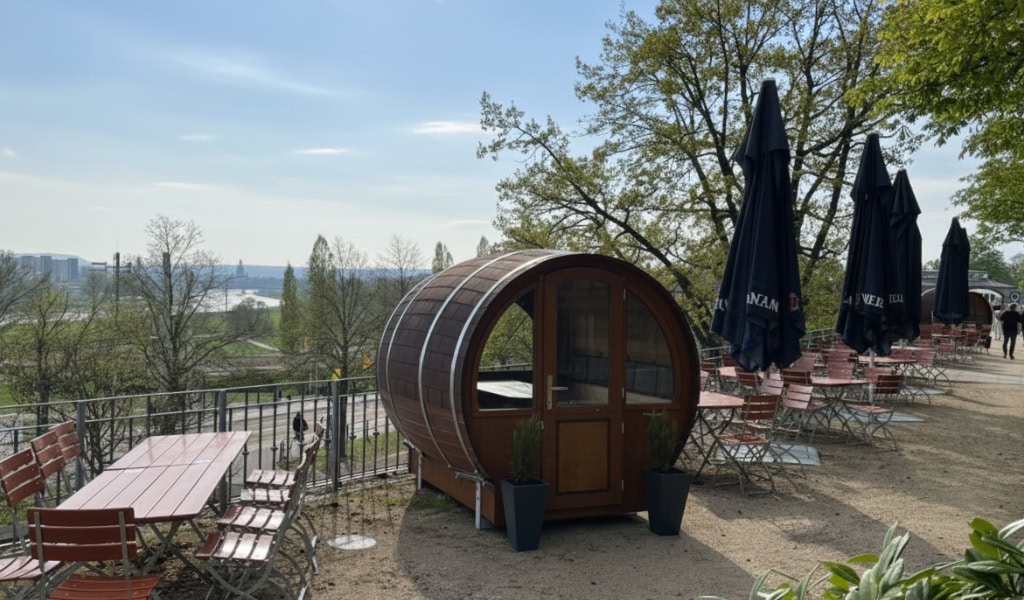 Sonnige Biergartenterrasse mit fassförmigem Holzhäuschen, leeren Tischen, Sonnenschirmen und Blick über Flusslandschaft.