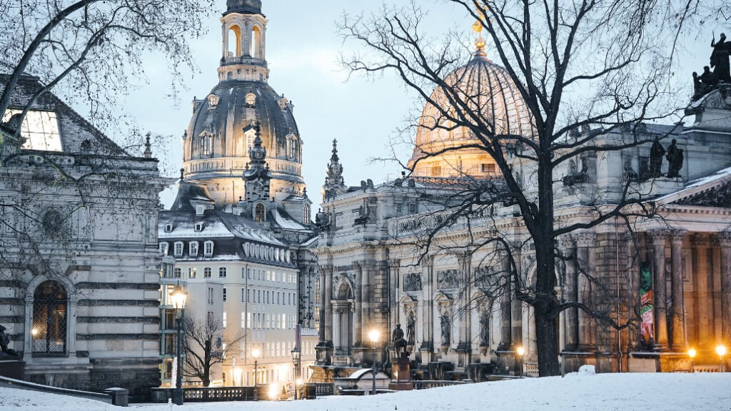 Winterlicher Blick auf die Dresdner Altstadt mit der Frauenkirche und der Kuppel der Kunstakademie im Abendlicht. Schnee bedeckt den Boden, während die historischen Gebäude stimmungsvoll beleuchtet sind.