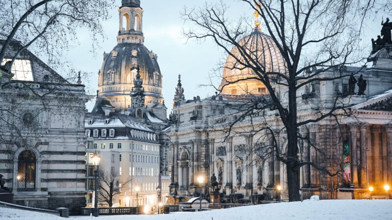 Winterlicher Blick auf die Dresdner Altstadt mit der Frauenkirche und der Kuppel der Kunstakademie im Abendlicht. Schnee bedeckt den Boden, während die historischen Gebäude stimmungsvoll beleuchtet sind.