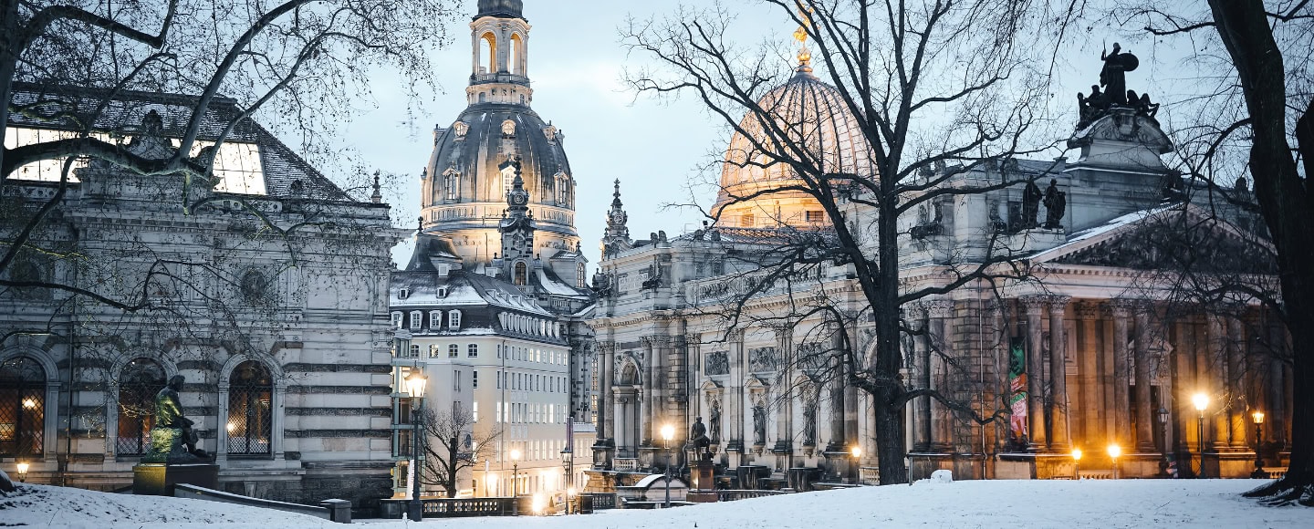 Winterlicher Blick auf die Dresdner Altstadt mit der Frauenkirche und der Kuppel der Kunstakademie im Abendlicht. Schnee bedeckt den Boden, während die historischen Gebäude stimmungsvoll beleuchtet sind.