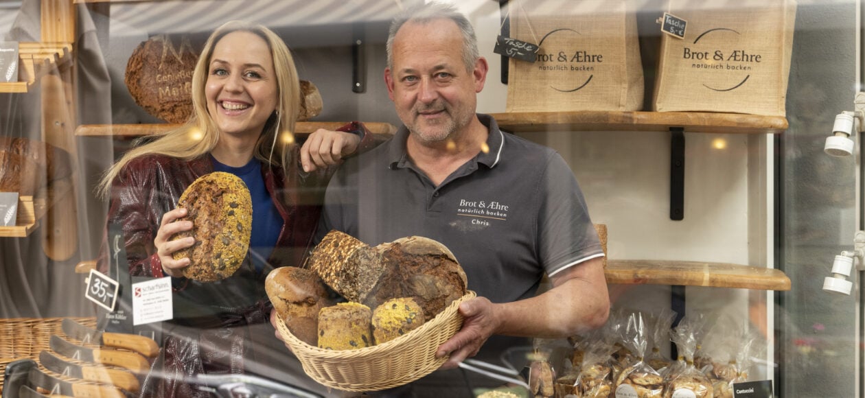Henriette Fee-Grützen steht neben Brotsommelier Chris Jentsch in seiner Bäckereii hinter einem Brotkorb voller Brötchen und Brote.