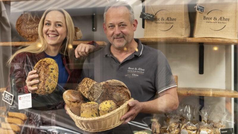 Henriette Fee-Grützen steht neben Brotsommelier Chris Jentsch in seiner Bäckereii hinter einem Brotkorb voller Brötchen und Brote.
