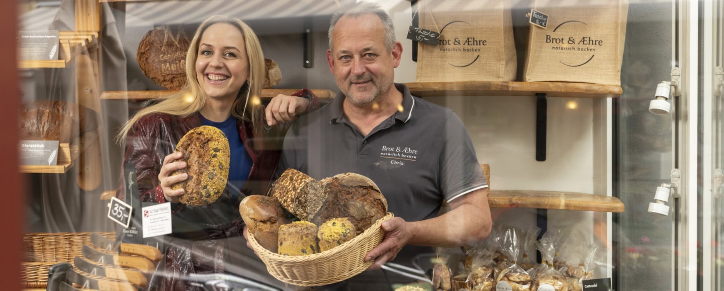 Henriette Fee-Grützen steht neben Brotsommelier Chris Jentsch in seiner Bäckereii hinter einem Brotkorb voller Brötchen und Brote.