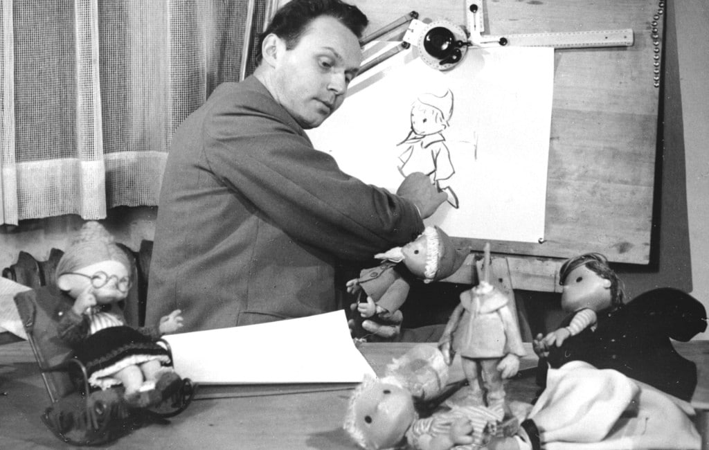 Historisches Schwarzweißfoto: Animator zeichnet eine Figur, umgeben von Puppen und Werkzeug – Einblick in analoge Produktion.