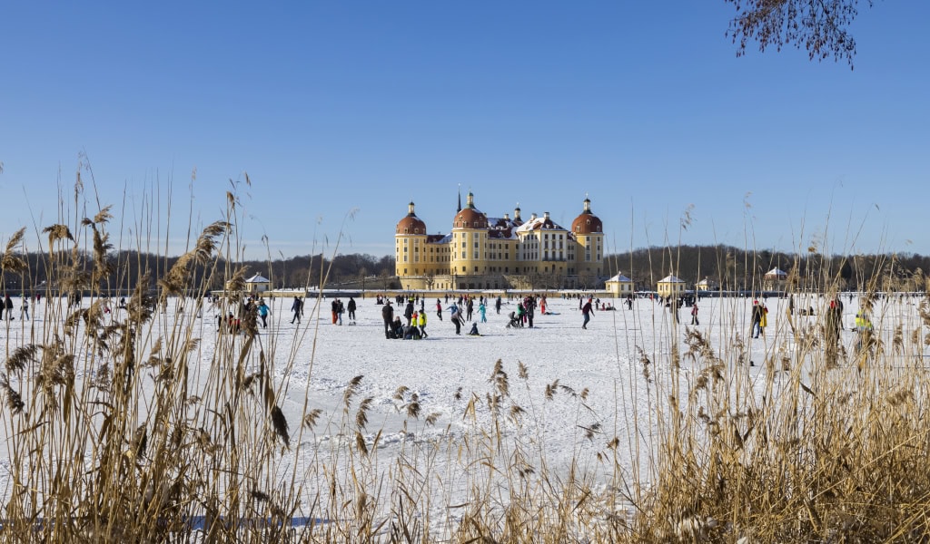 Blick auf die zugefrorenen Teich mit Spaziergängern und Eisläufern vor Schloss Moritzburg