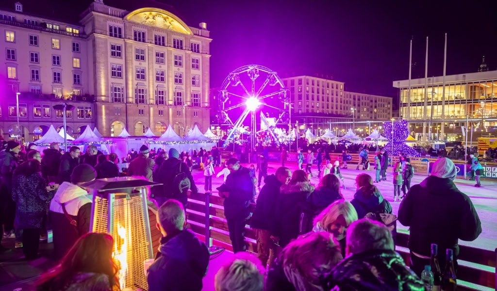Auf einer bunt angestrahlten Eisfläche am Dresdner Altmarkt fahren Menschen Schlittschuh. Im HIntergrund ist ein Riesenrad