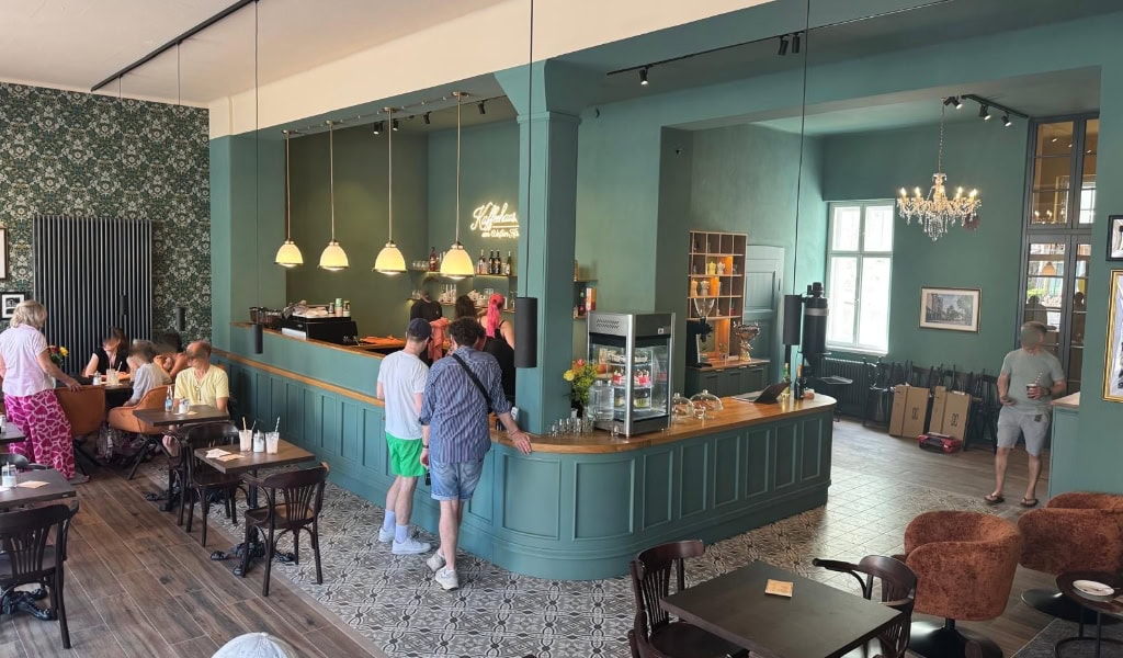 Innenraum des Kaffeehauses am Weißen Hirsch mit türkisgrüner Theke, gemütlichen Sitzplätzen und Gästen.
