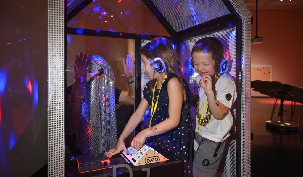 Zwei Kinder tanzen mit Kopfhörern in einer beleuchteten Silent-Disco-Kabine und bedienen gemeinsam ein Mischpult.