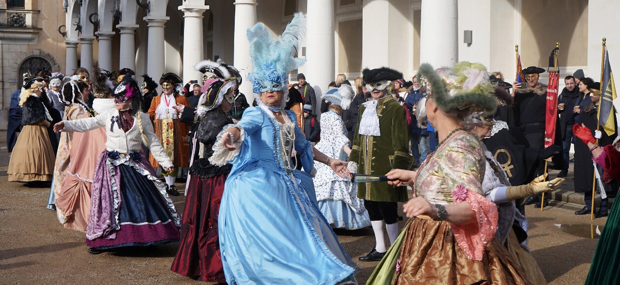 Mehrere Teilnehmerinnen und Teilnemer des Elbvenezianischen Carnevals in Dresden tanzen gemeinsam in farbenfohen, kunstvollen Kleidern und Masken im Barock-Stil.