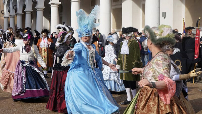 Mehrere Teilnehmerinnen und Teilnemer des Elbvenezianischen Carnevals in Dresden tanzen gemeinsam in farbenfohen, kunstvollen Kleidern und Masken im Barock-Stil.