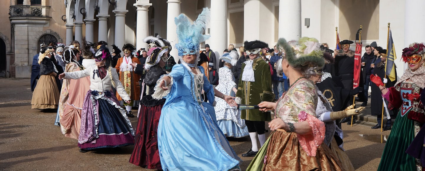Mehrere Teilnehmerinnen und Teilnemer des Elbvenezianischen Carnevals in Dresden tanzen gemeinsam in farbenfohen, kunstvollen Kleidern und Masken im Barock-Stil.