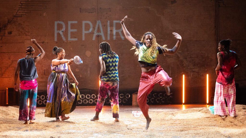 Fünf Tänzer:innen in bunten Outfits bewegen sich auf sandbedeckter Bühne, eine Person springt hoch beim Tanzfestival.