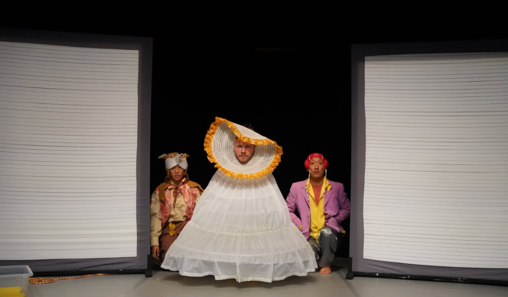 Figur im großen weißen Kleid mit orangener Kragenform zwischen zwei knienden Darstellenden auf Bühne des Tanzfestivals.