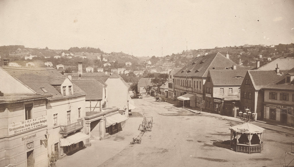 Weite, helle Aufnahme eines städtischen Platzes mit Geschäften und Markisen. Wenige Karren, kaum Menschen; klare Sicht auf die Hänge dahinter.