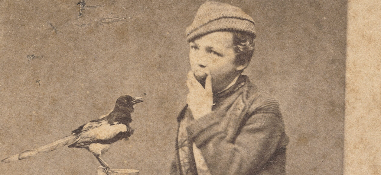 Sepiaporträt: Junge Person mit Mütze blickt zur Kamera; eine Elster sitzt auf einem Holzständer daneben.