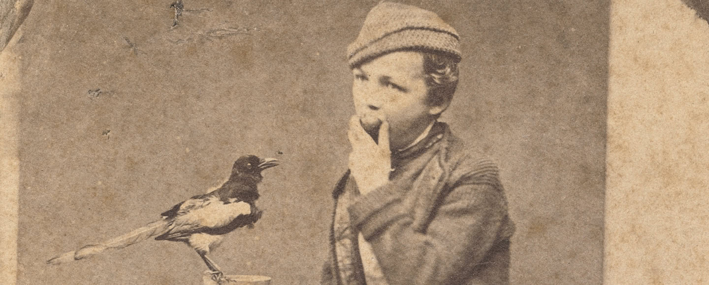 Sepiaporträt: Junge Person mit Mütze blickt zur Kamera; eine Elster sitzt auf einem Holzständer daneben.