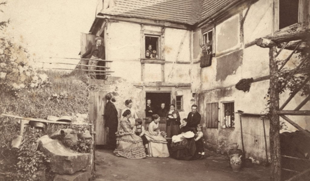 Historische Hofszene vor einem Fachwerkhaus: Mehrere Personen sitzen und stehen in Gruppen, einige blicken aus Fenstern.