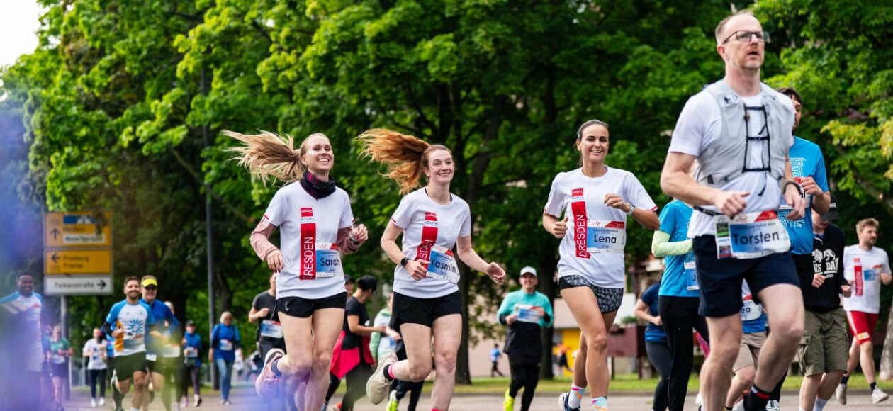 Mehrere Läuferinnen und Läufer nehmen an einer Straßenlauf-Veranstaltung in Dresden teil und joggen auf einer von grünen Bäumen gesäumten Strecke durch die Stadt.