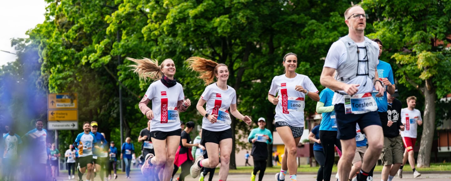 Mehrere Läuferinnen und Läufer nehmen an einer Straßenlauf-Veranstaltung in Dresden teil und joggen auf einer von grünen Bäumen gesäumten Strecke durch die Stadt.