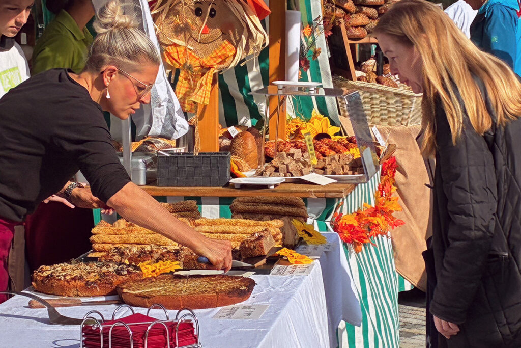 Herbstlich geschmückter Marktstand mit Kuchen und Brotsorten, über den sich eine Kundin beugt. 